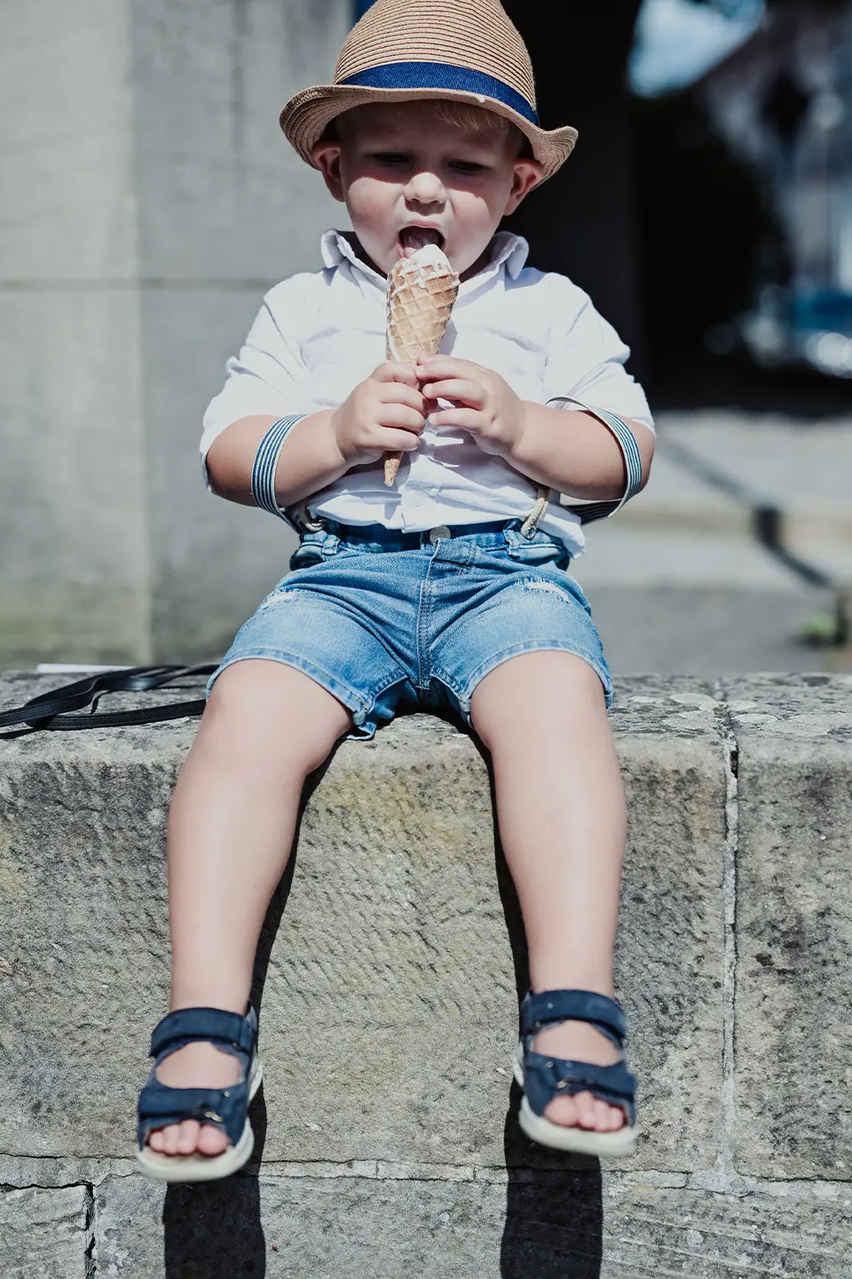 kleiner Junge mit Hut sitzt auf einer Mauer und ißt ein Eis.