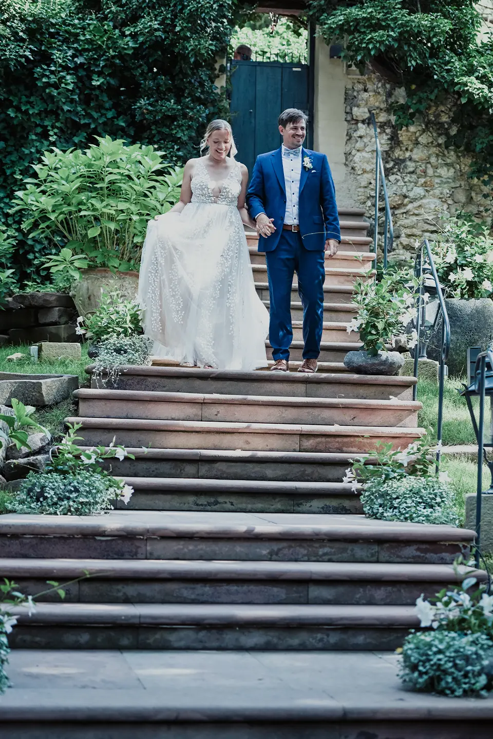 Foto eins Hochzeitpaares das lachend eine Treppe hinunterläuft mit schönen unscharfen Blättern im Vordergrund.