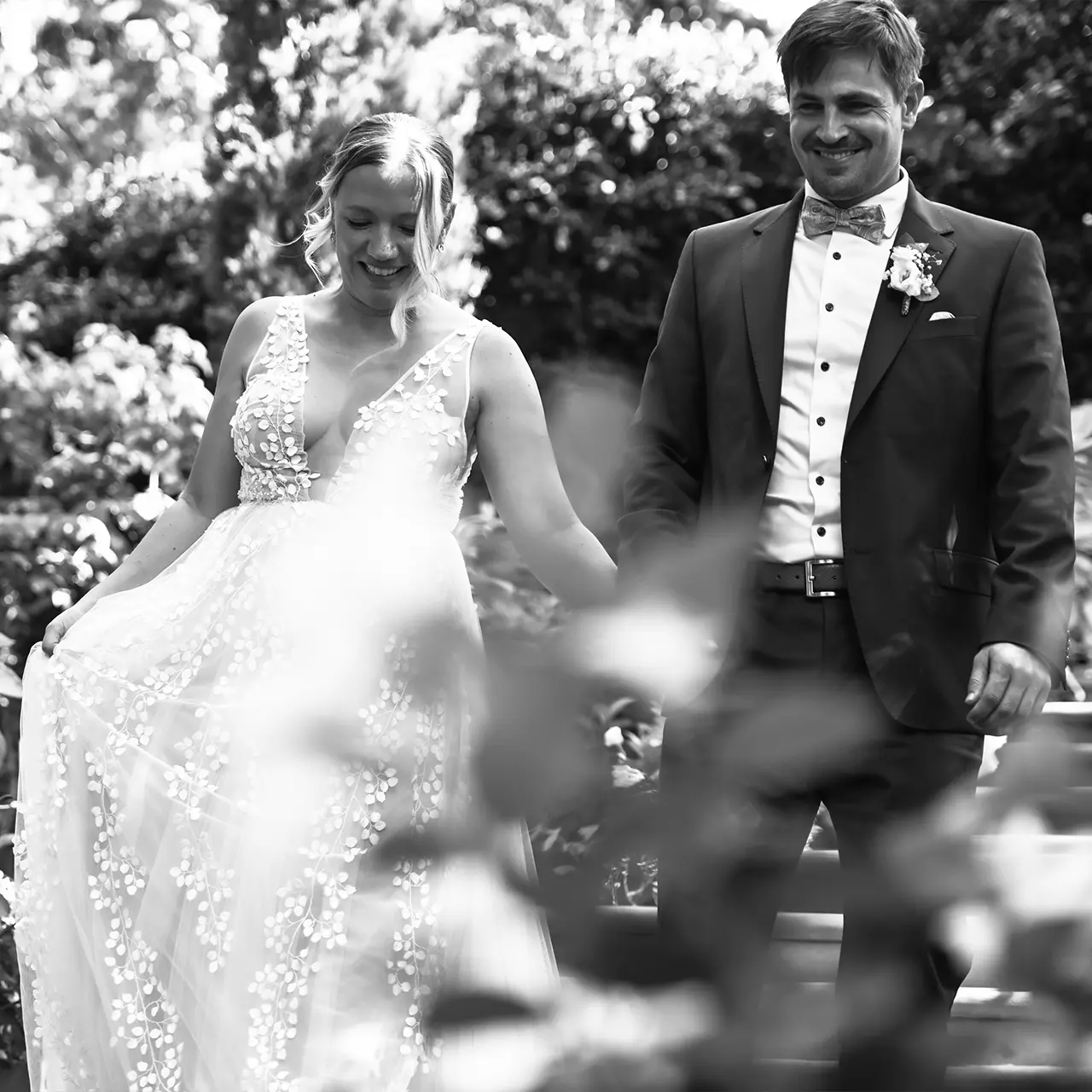 Schwarz-weiß Foto eins Hochzeitpaares das lachend eine Treppe hinunterläuft mit schönen unscharfen Blättern im Vordergrund.