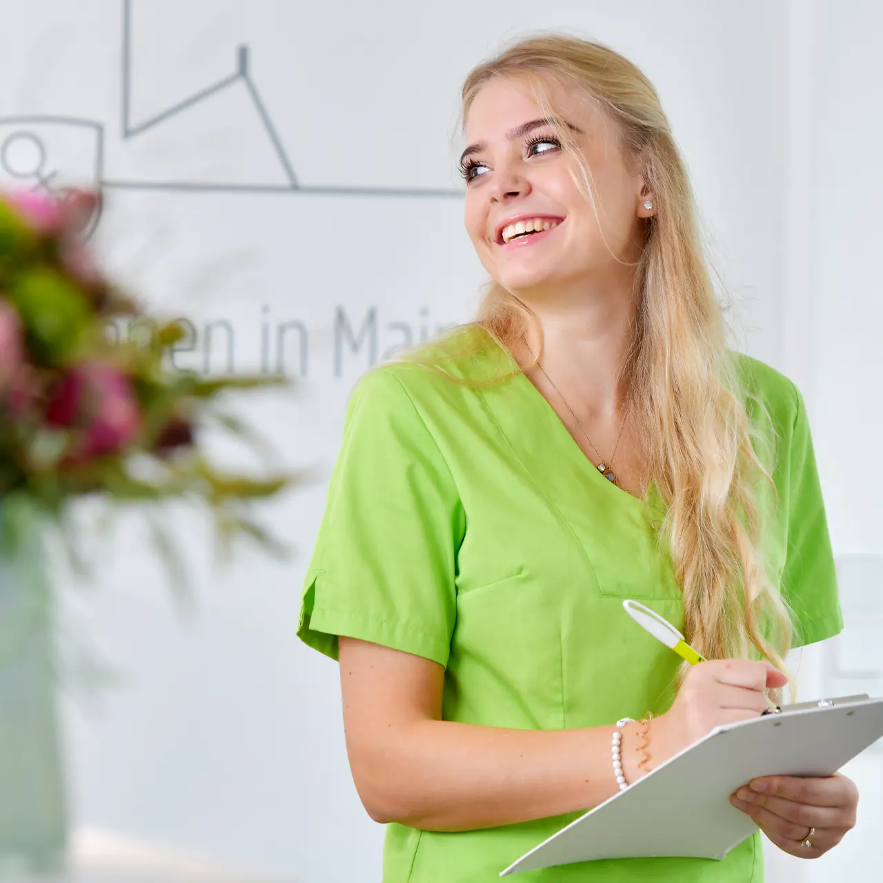 Personalfoto einer jungen Frau im grünen Kittel, Mitarbeiterin einer gynäkologischen Praxis, mit rosa Blumen im Hintergrund.
