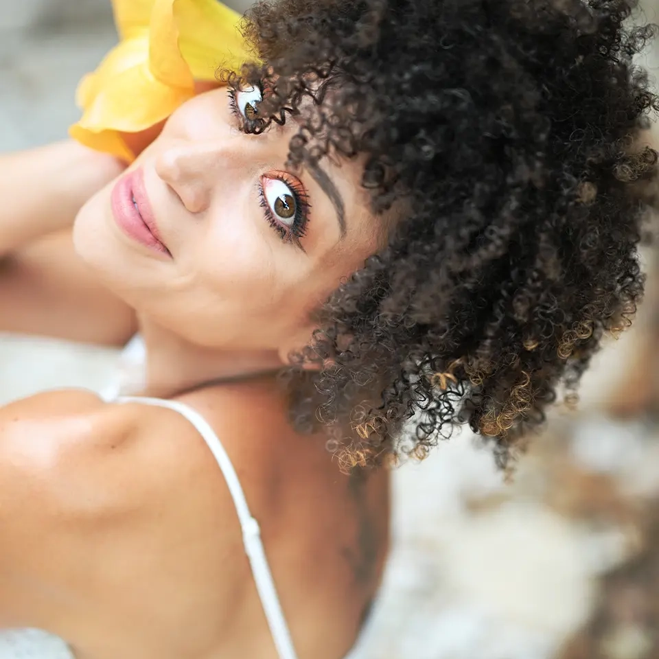 Portrait einer Frau mit Hibiskusblüte und wilden Locken, die nach oben in die Kamera schaut.